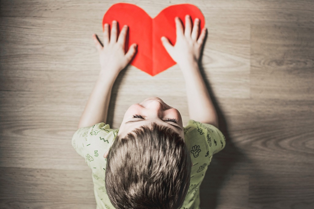 Looking from above, you can see the head of a young child with blond hair on floor with hands on a large red  paper heart