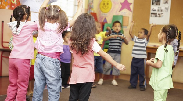 8-12  children of both genders (ages 4-5)  in a standing circle in a classroom doing an activity or dance with arms in the air.