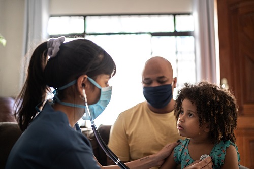 Home health visitor with mask checking vitals with parent wearing mask in background.