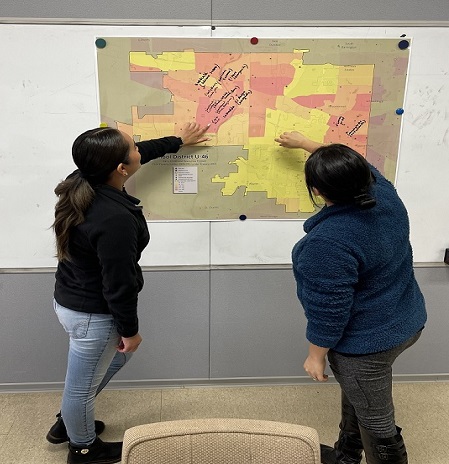 Two Latina women writing on a large map pinned to a wall. The map shows their school district with different color census tracts corresponding to areas of higher poverty.