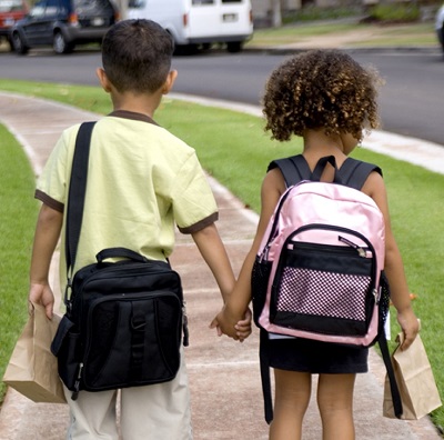 young boy and girl holding hands and walking to school with backpacks on (showing their backs)