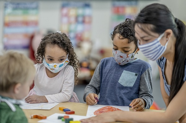 Multi-ethnic group of children coloring at a table while wearing protective face masks with teache