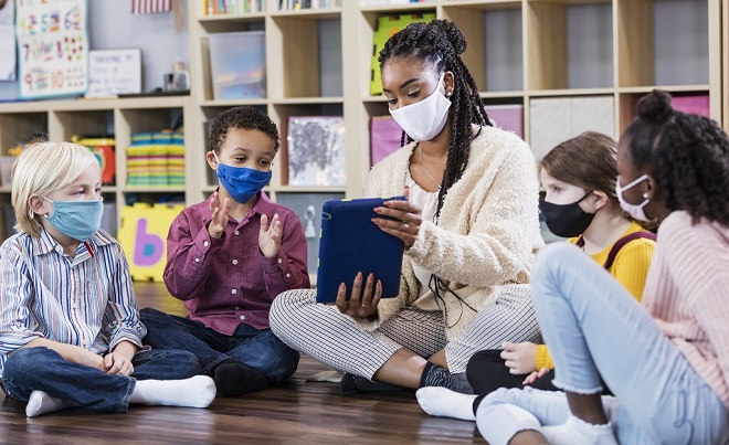 Teacher showing diverse students something on an Ipad while seated on the floor wearing masks.